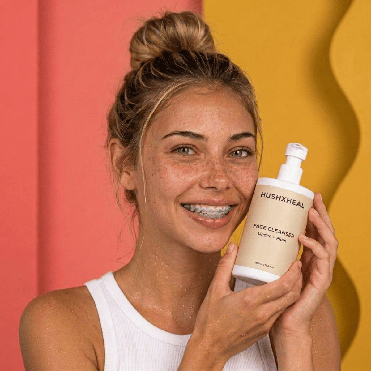 Smiling woman with freckles and hair in a messy bun holding HUSHXHEAL Linden + Plum Face Cleanser beside her face during her skincare routine.