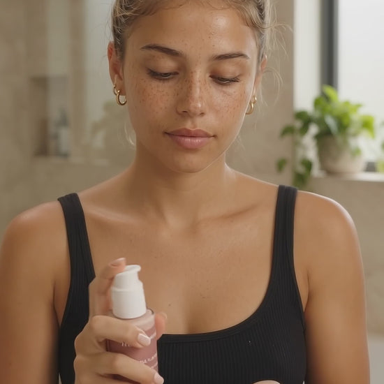 Young woman with freckles and a messy blonde bun wears a black tank in a naturally lit bathroom. She pumps HUSHXHEAL Renewal Serum into her palm, rubs her hands together, and gently presses it onto her face.