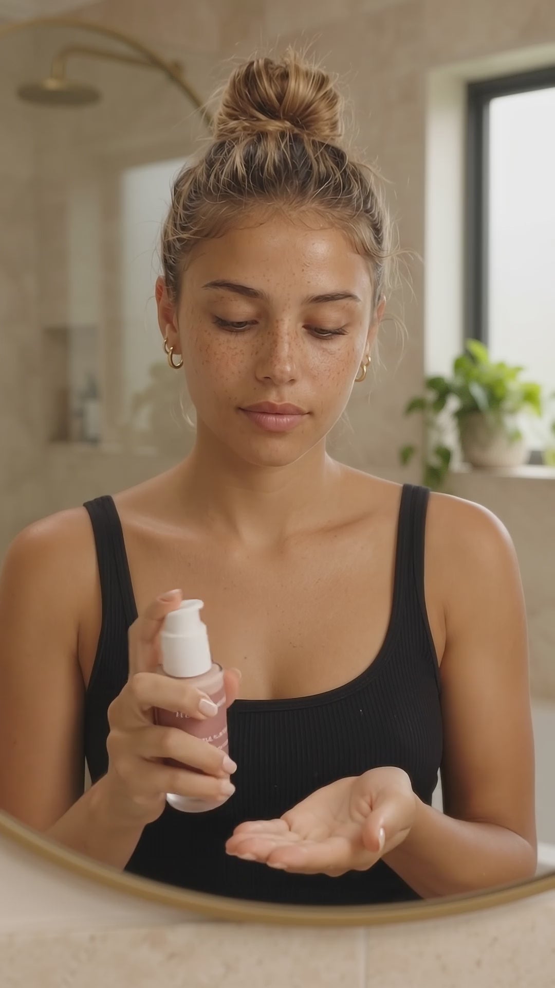 Young woman with freckles and a messy blonde bun wears a black tank in a naturally lit bathroom. She pumps HUSHXHEAL Renewal Serum into her palm, rubs her hands together, and gently presses it onto her face.